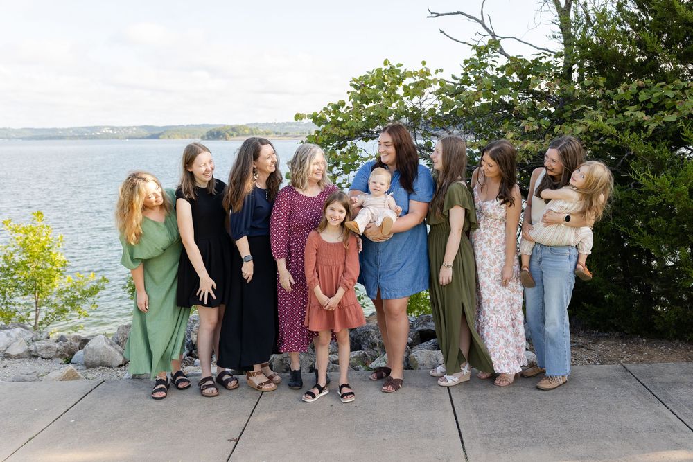 Group of women and girls posing outdoors near a waterfront with lush greenery and a scenic lake in the background.