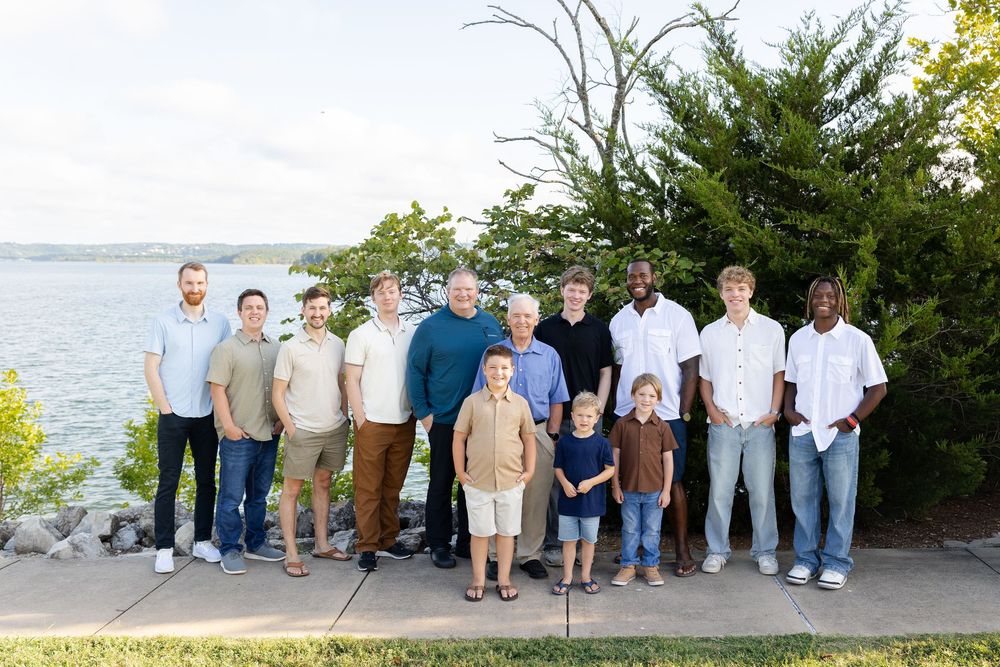 Group of men and boys posing outdoors near a lakeside with trees and rocky shoreline in background.