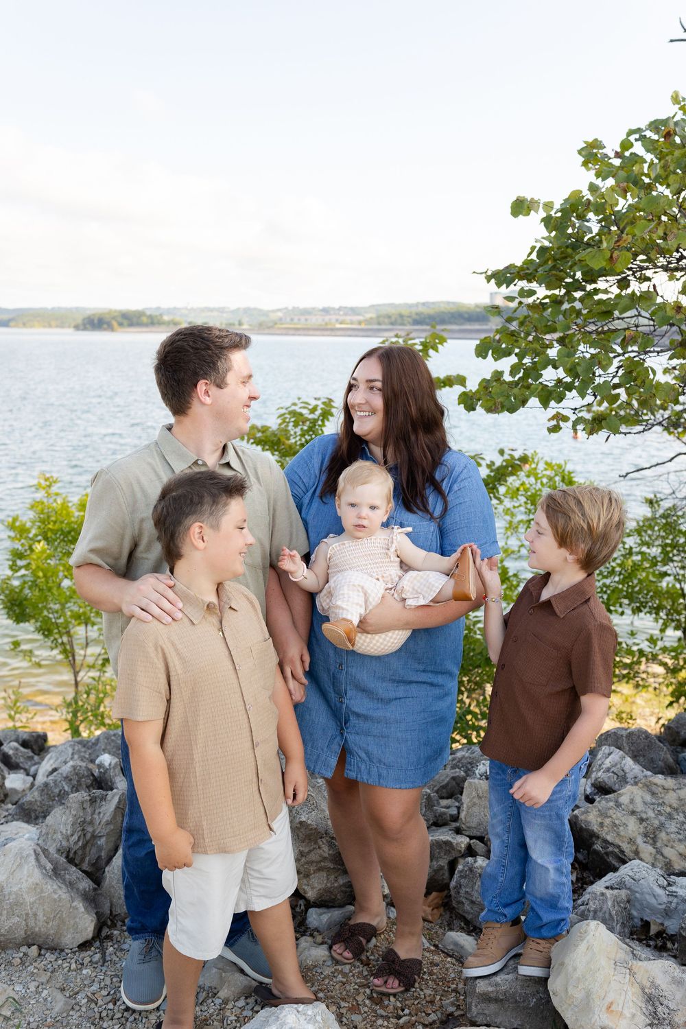 Happy family of five posing on rocky lakeside, parents with three children including baby, surrounded by lush greenery.