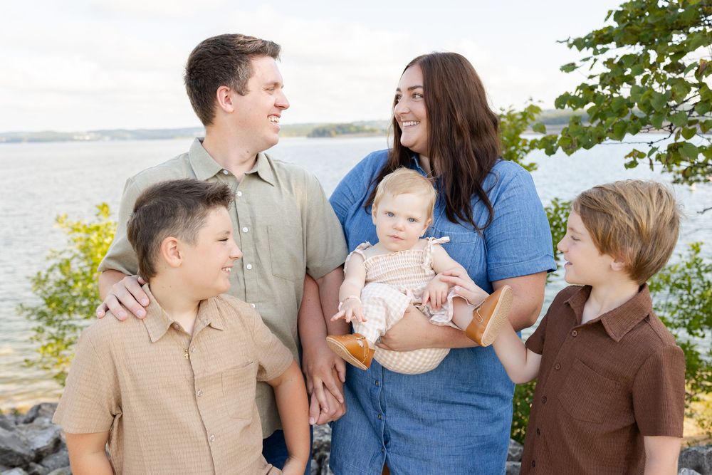 Family of five posing outdoors near a lake, parents with three children including a baby, surrounded by lush greenery.