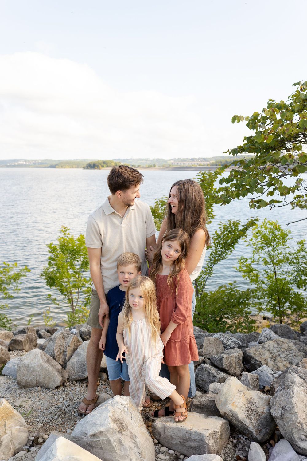 Family of five posing on rocky lakeside shore surrounded by green trees on a sunny day.