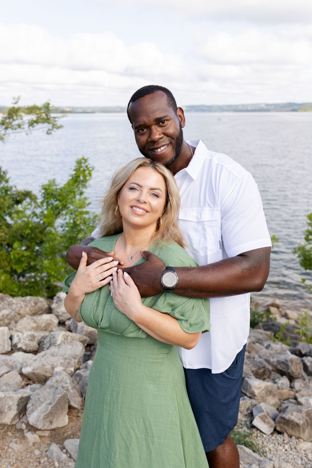 A smiling interracial couple embraces outdoors near a rocky shoreline with trees and water in the background.