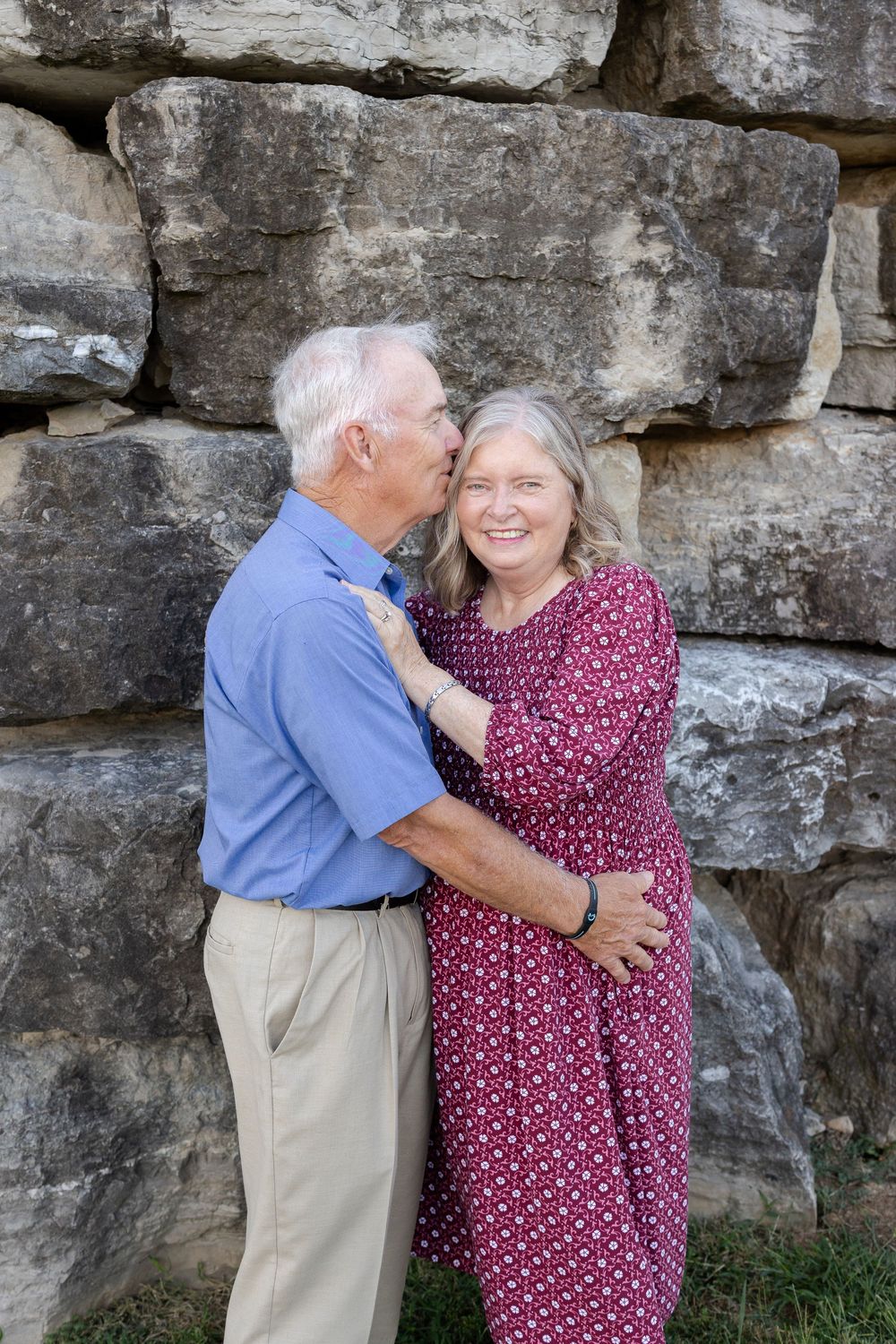 Elderly couple embracing and sharing a kiss in front of a rustic stone wall outdoors.