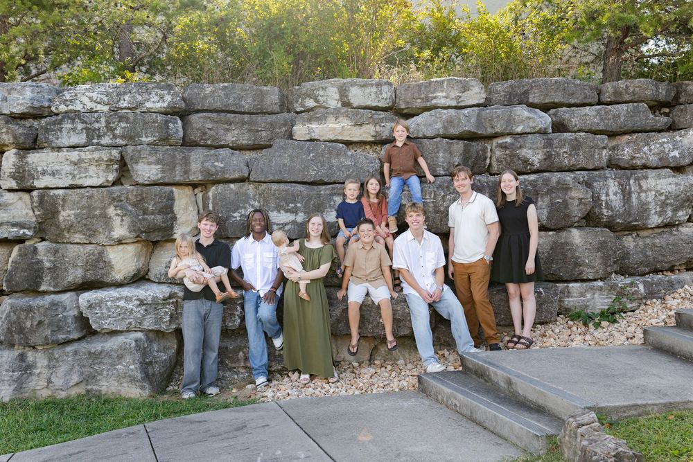 Large family group portrait posed in front of a rustic stone wall surrounded by lush green trees outdoors.