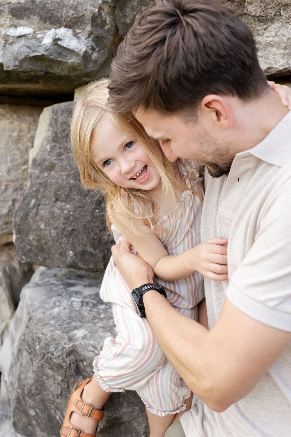 A father lovingly embraces his smiling blonde daughter against a stone wall backdrop.