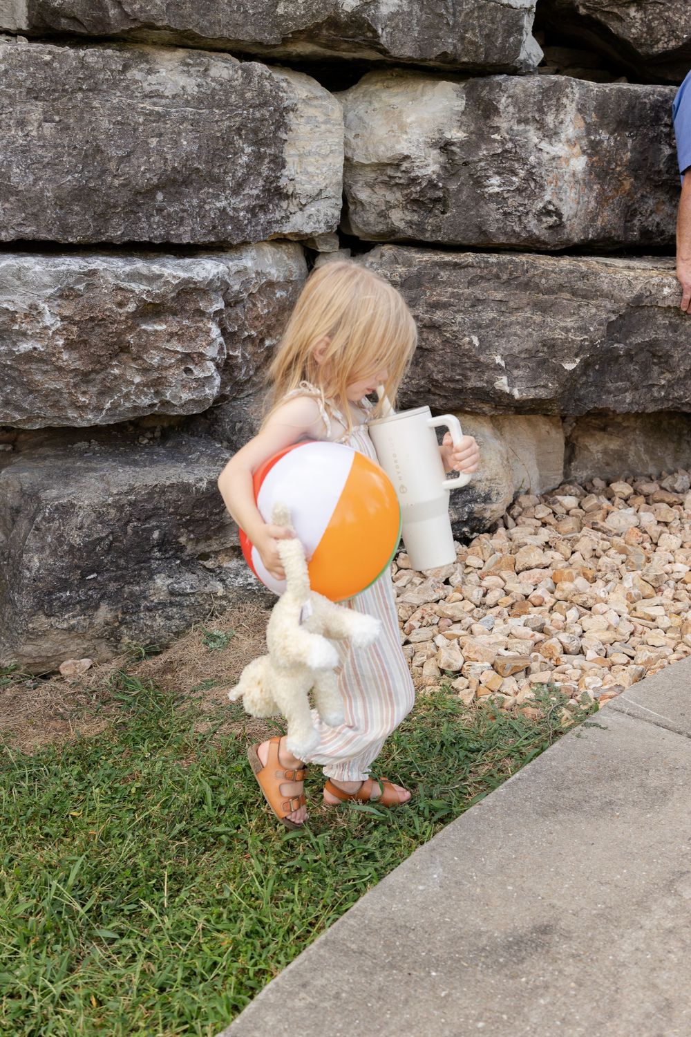Toddler girl carrying a beach ball, stuffed animal, and cup near a stone wall outdoors.