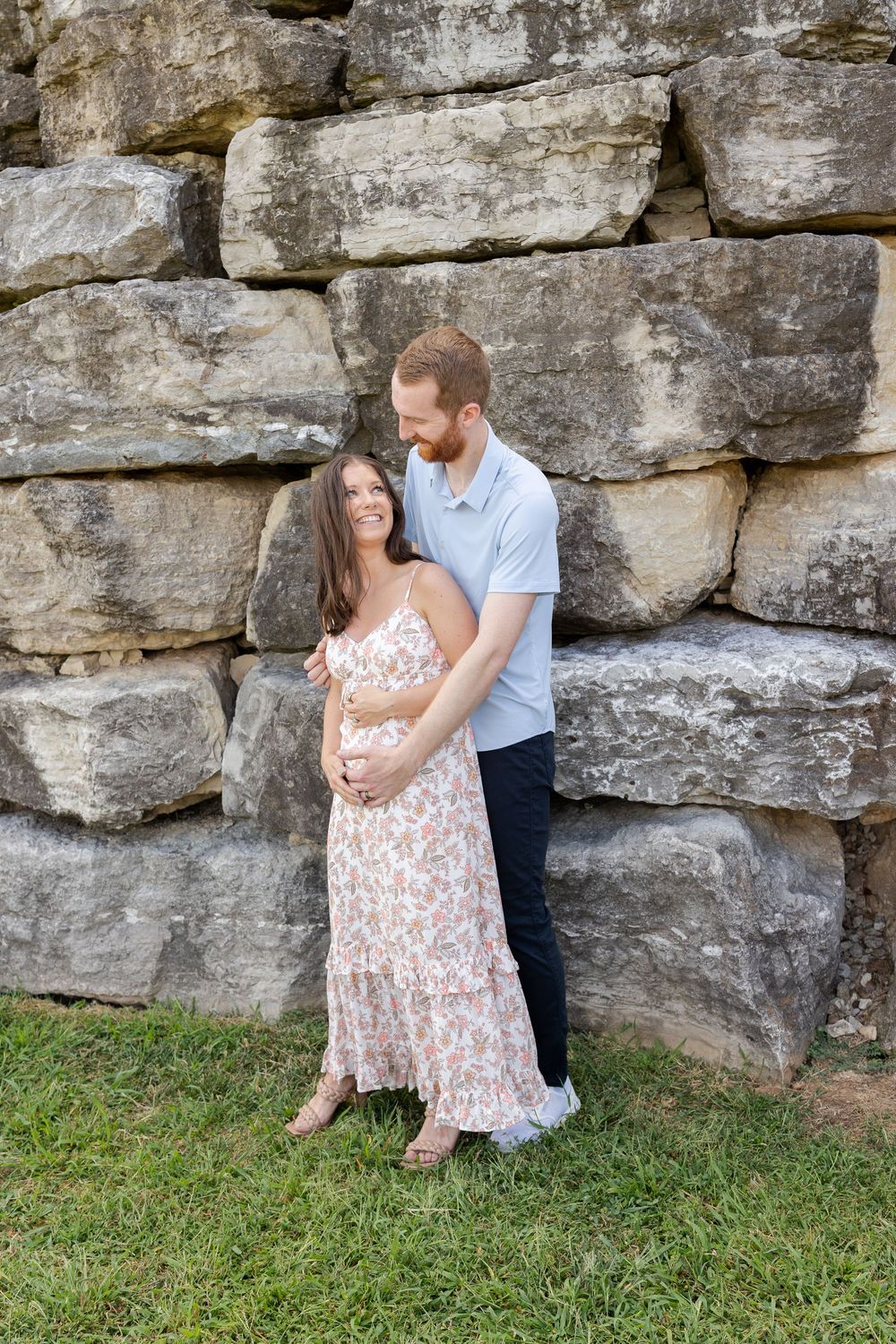 Couple embracing in front of a stone wall outdoors, woman in floral dress with man in blue shirt holding her.