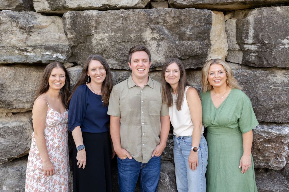 Five smiling adults posing together in front of a rustic stone wall outdoors.