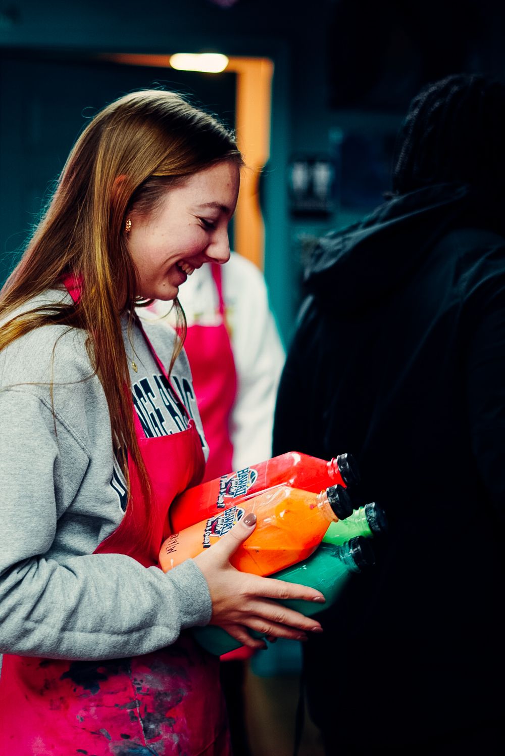 Smiling young woman in pink apron holding colorful bottles at an event.
