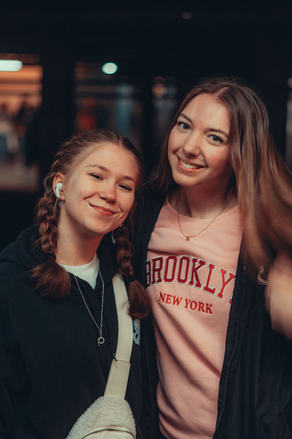 Two smiling young women posing together, one wearing a pink Brooklyn New York sweatshirt, indoors at night.