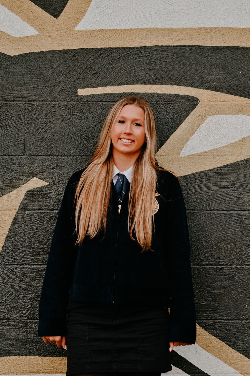 Smiling blonde teen in black FFA jacket and tie poses against a painted mural wall outdoors.
