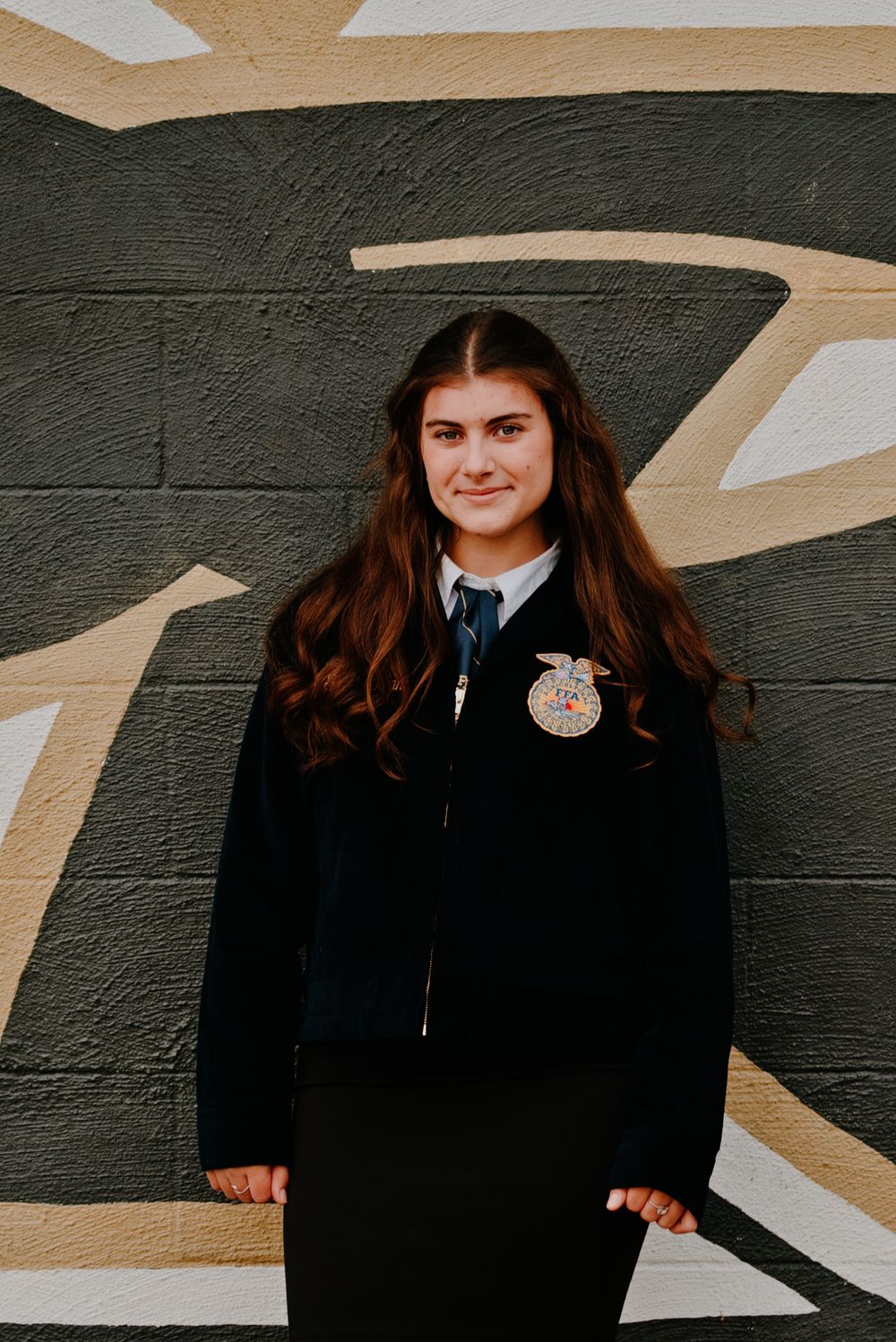 Young woman in FFA official dress jacket with emblem poses smiling before a painted mural wall.