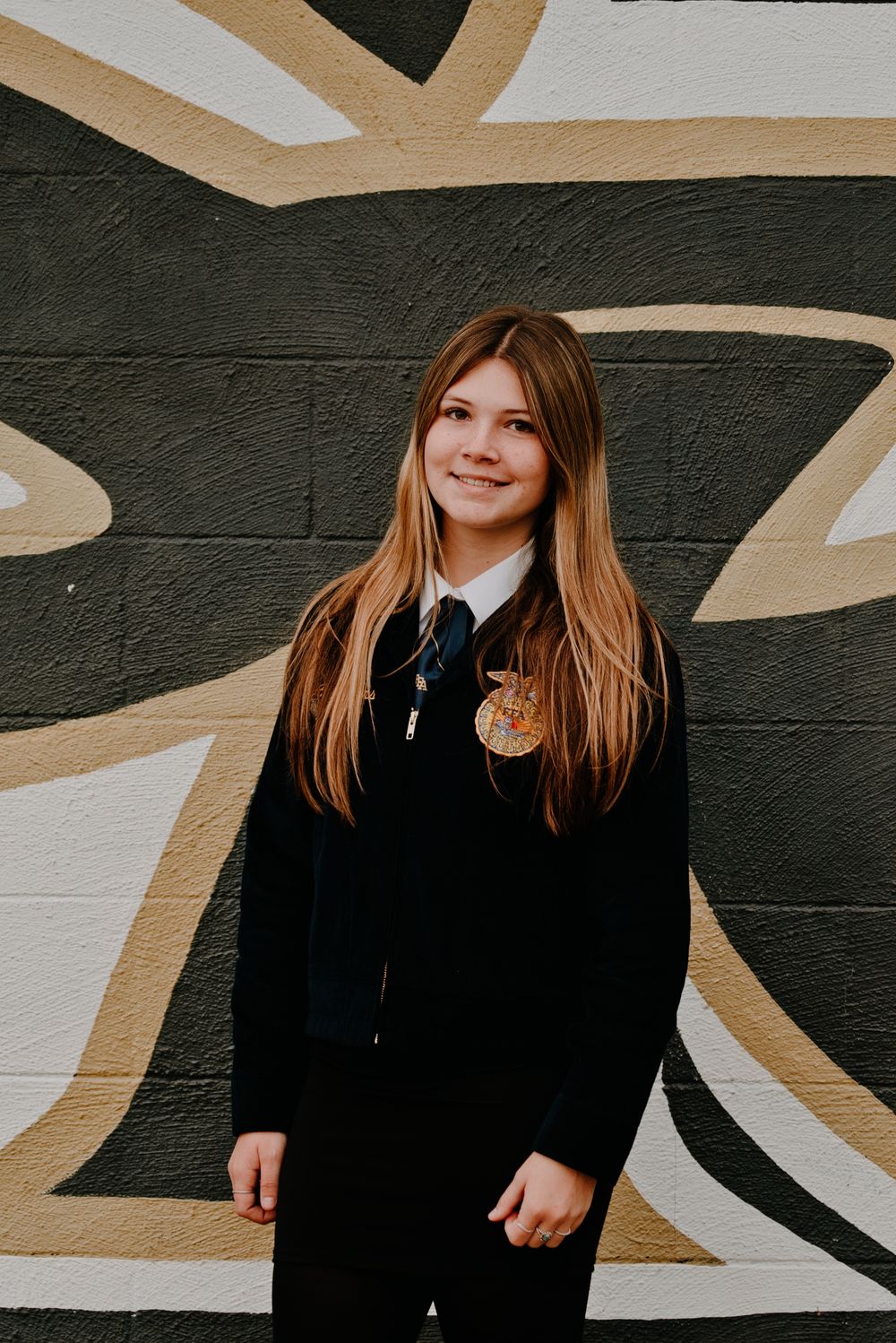 Young FFA member smiling in black jacket with emblem badge, standing before a painted mural wall.