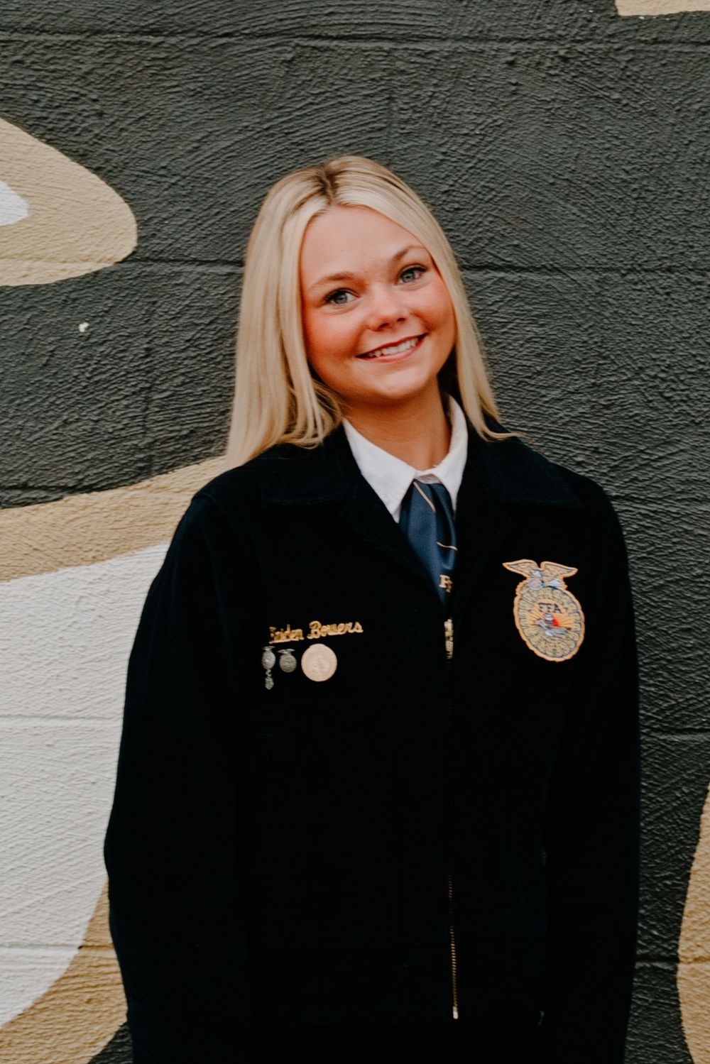 Young woman smiling in official FFA jacket with emblem badge, posing in front of a camouflage-painted wall.