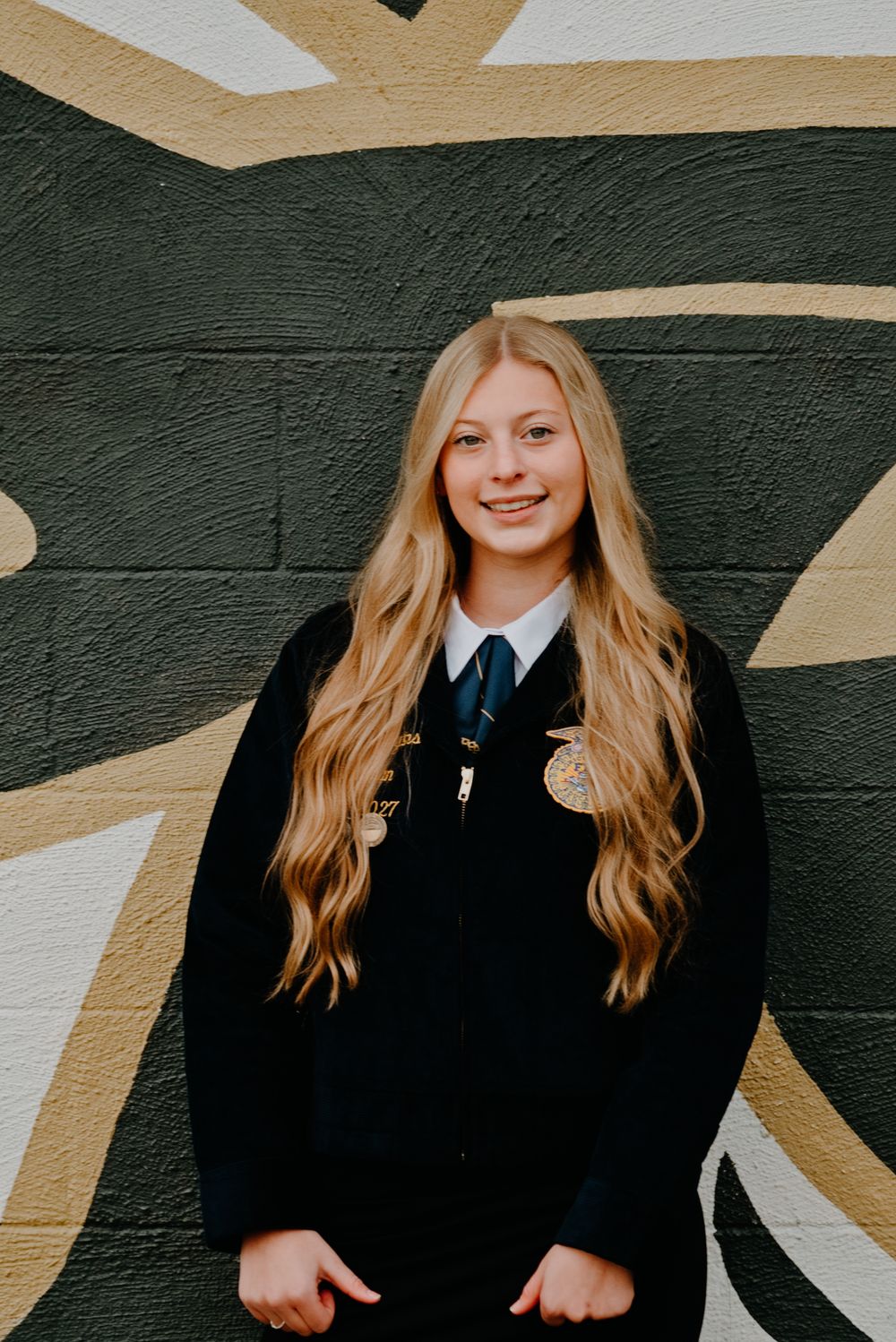 A smiling teenage girl with long blonde hair wearing a black FFA jacket and tie, posing in front of a painted brick wall.