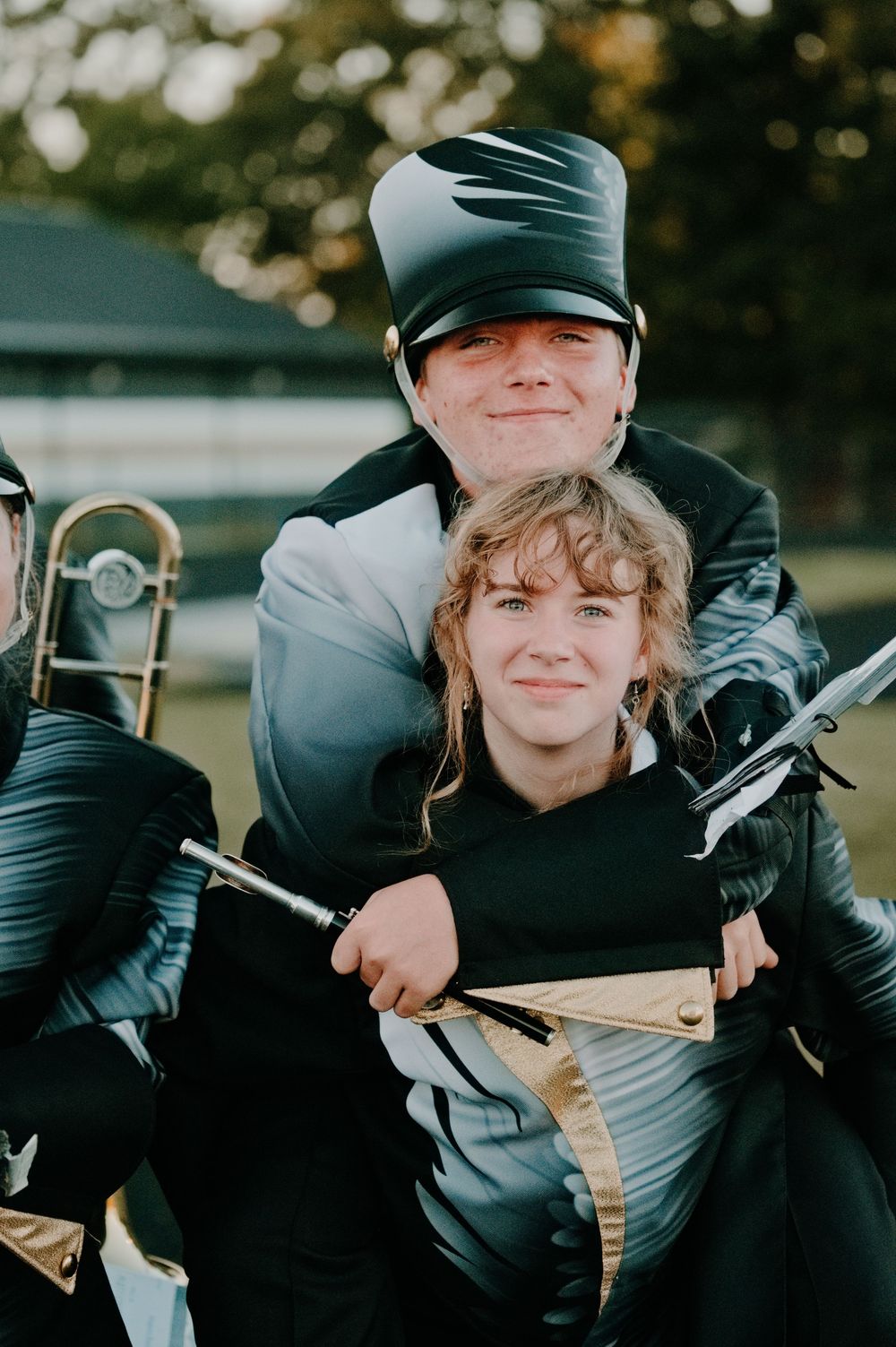 Two marching band members in black uniforms smile together outdoors, one holding a rifle prop.