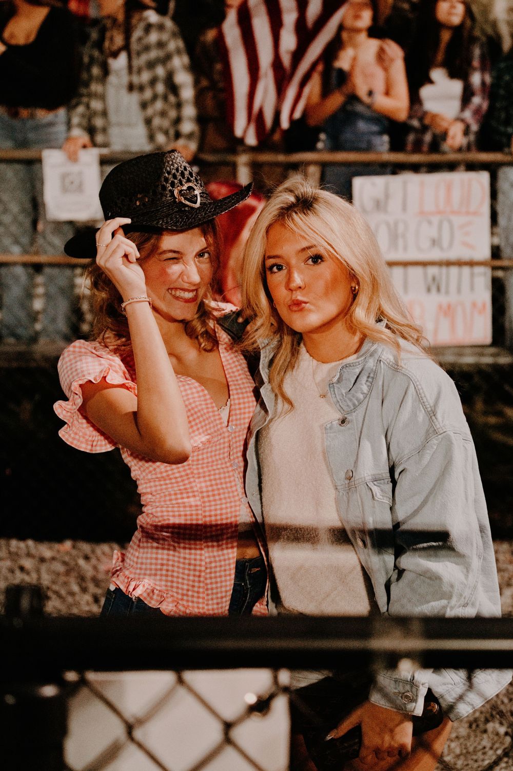 Two young women posing at a rodeo event, one wearing a black cowboy hat and pink gingham top.