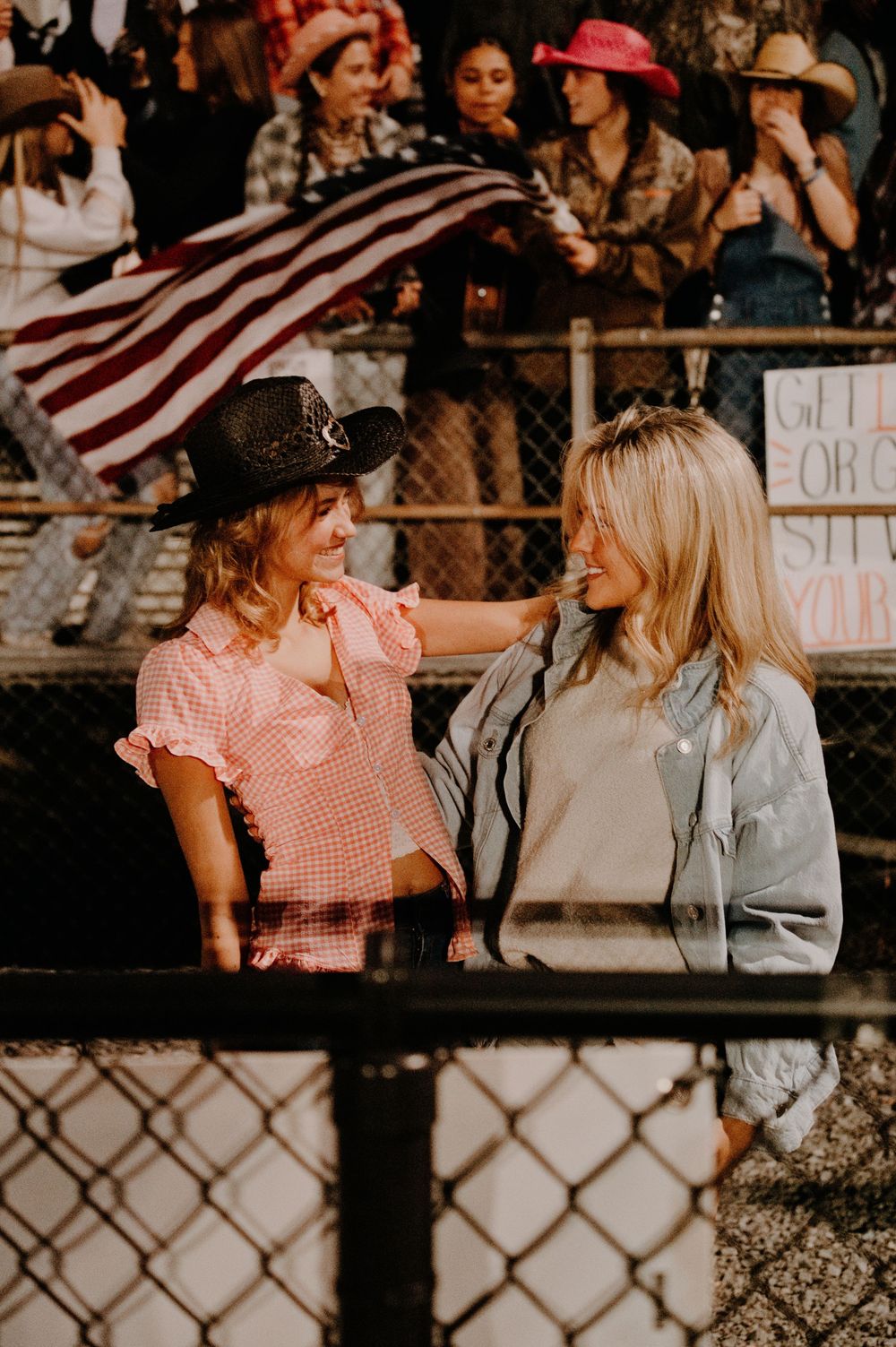 Two women in cowgirl attire smile at each other at a football game with an American flag and crowd behind them.