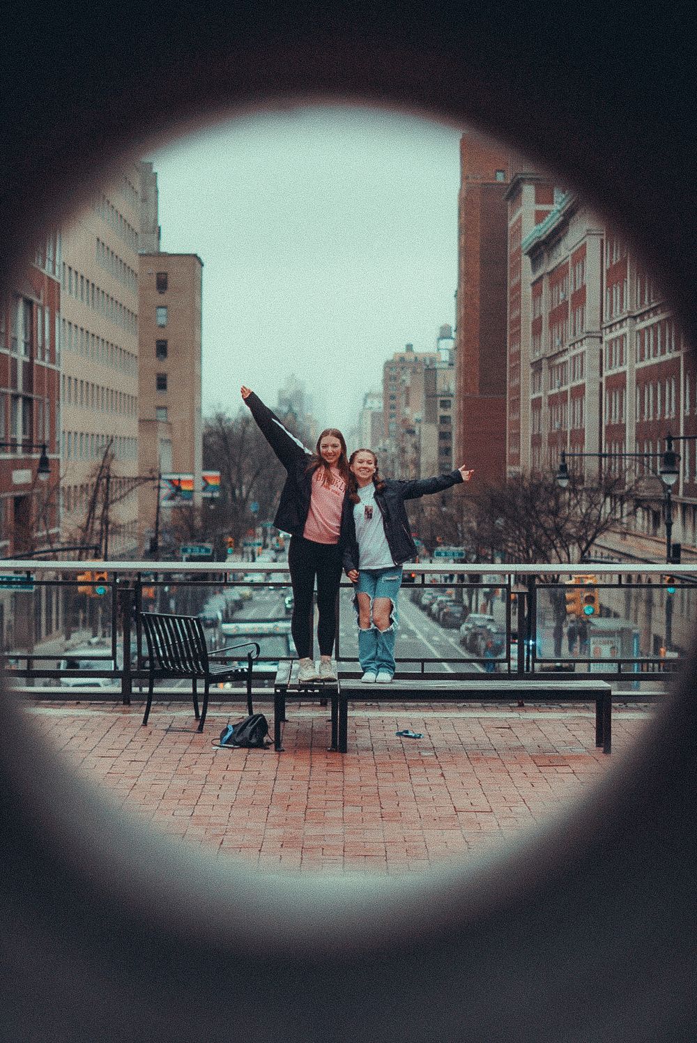 Two girls pose with arms outstretched on a city overlook, framed by a circular tunnel opening with urban buildings behind.