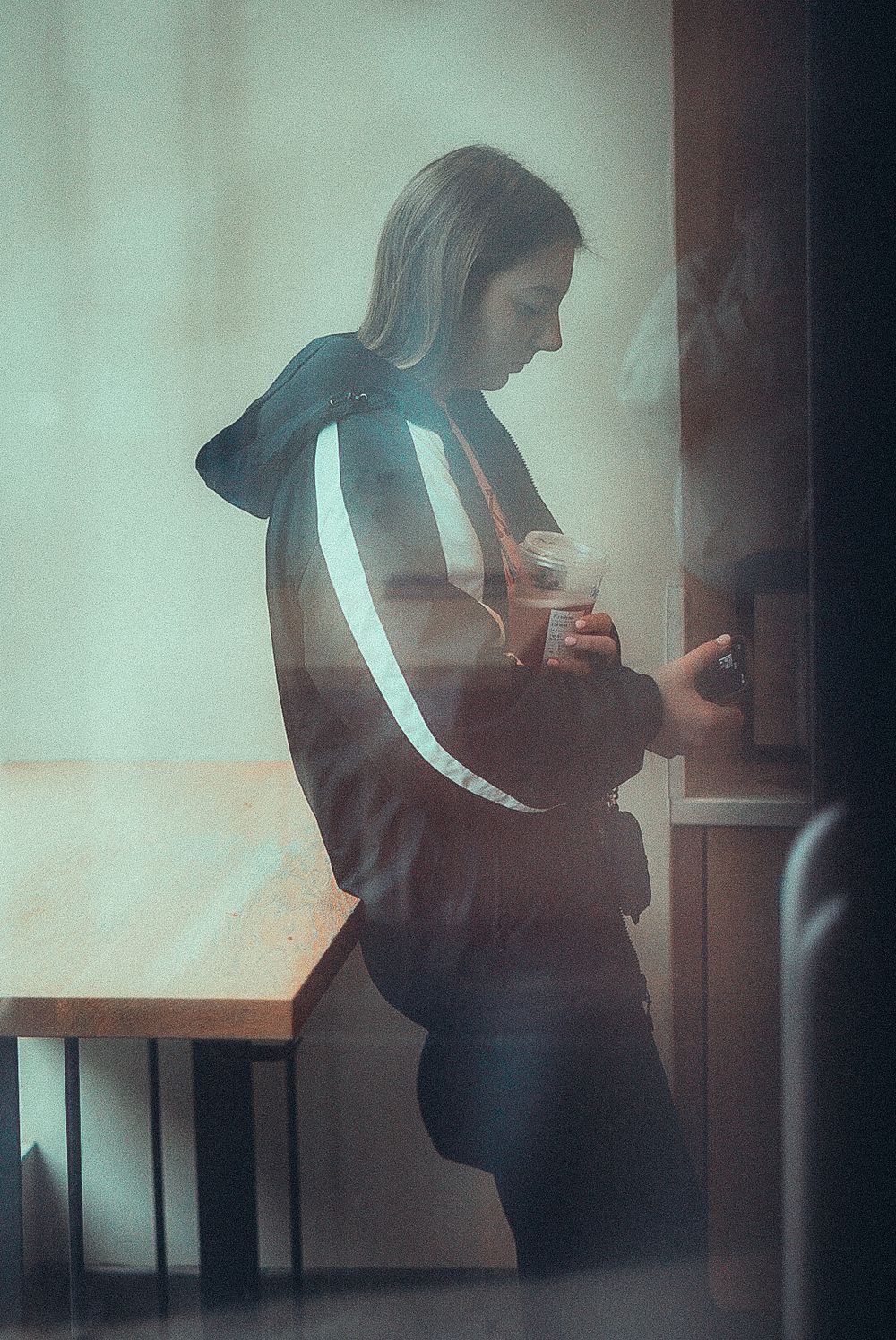 Person in a reflective stripe jacket holding a drink, photographed through glass in a moody, film-style portrait.
