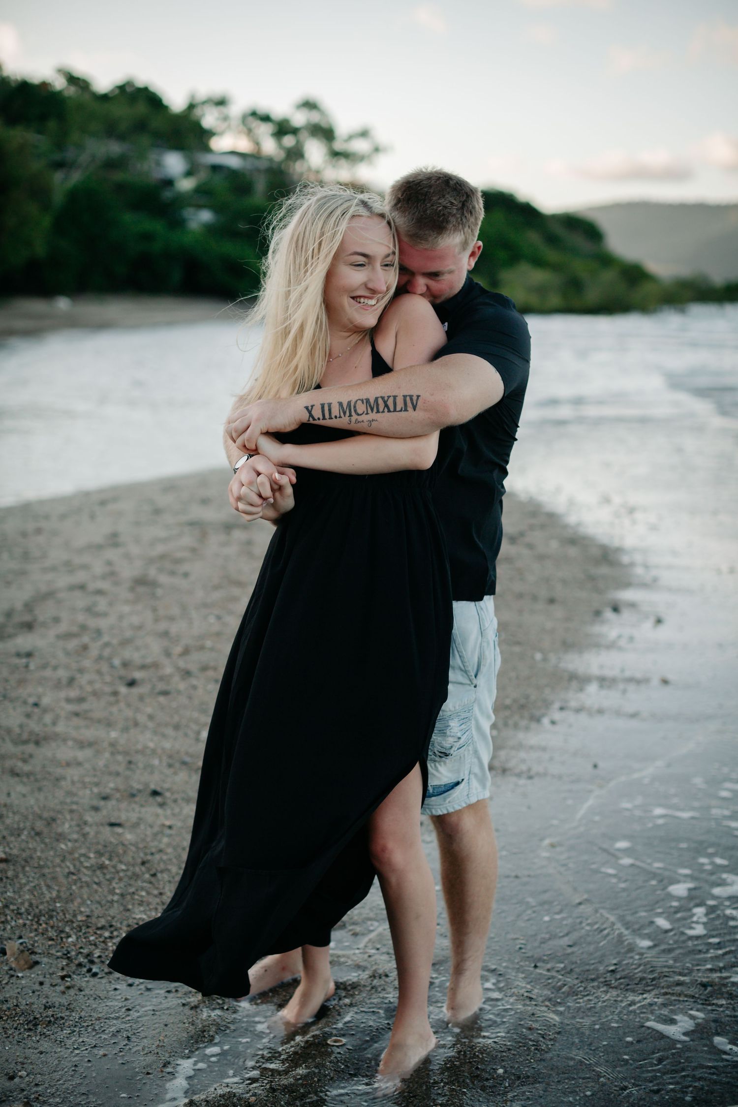 Couple embracing on a beach at dusk, woman in black dress with tattoo, man in black shirt and shorts.