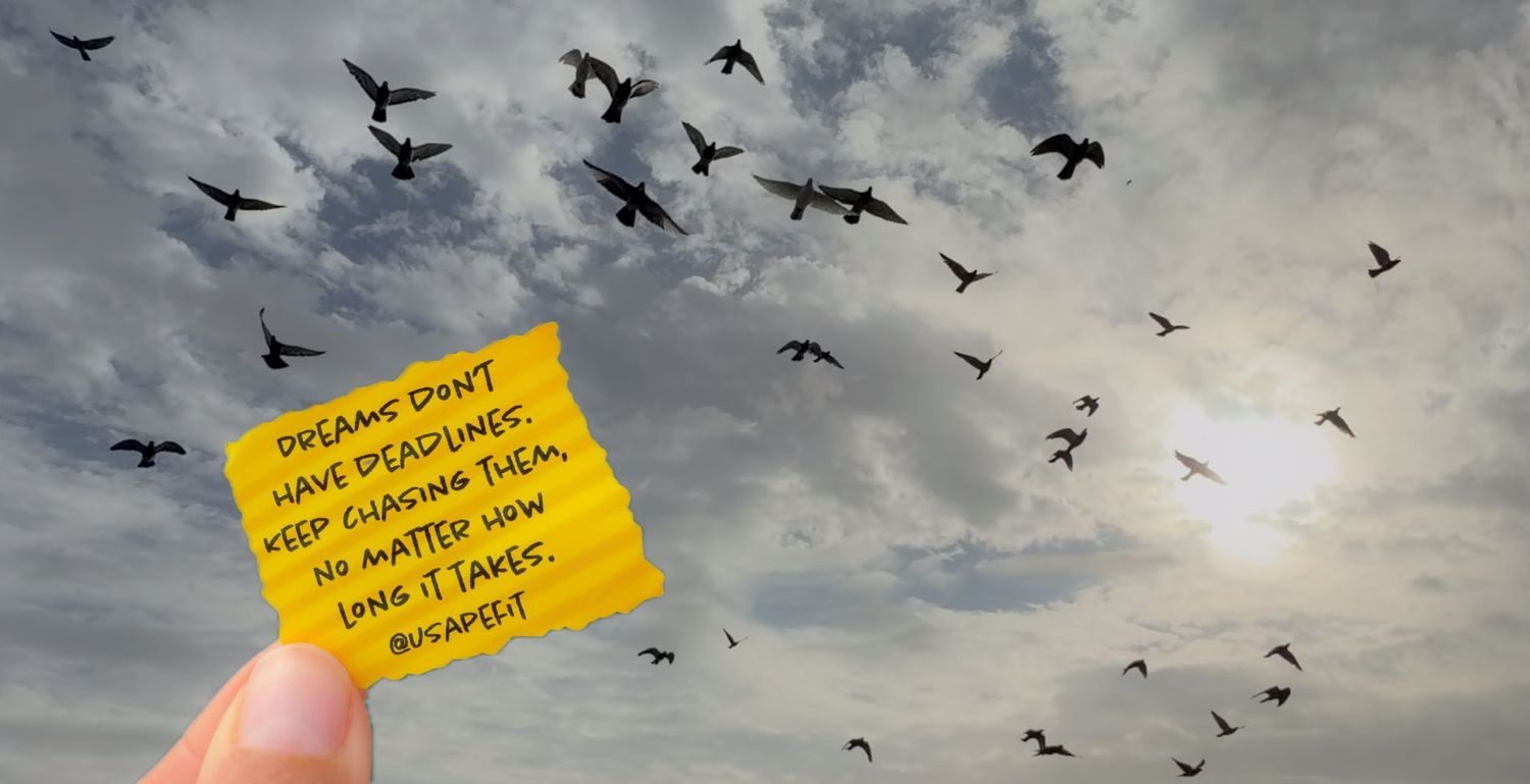 A hand holds a yellow sticky note reading dreams dont have deadlines against a dramatic sky filled with flying birds.