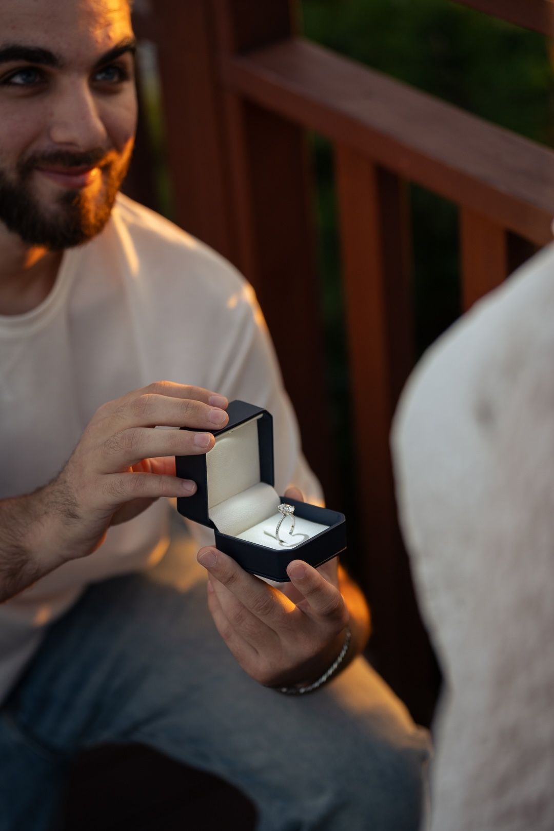 Smiling bearded man presenting an open ring box with a diamond ring during a romantic outdoor proposal at dusk.
