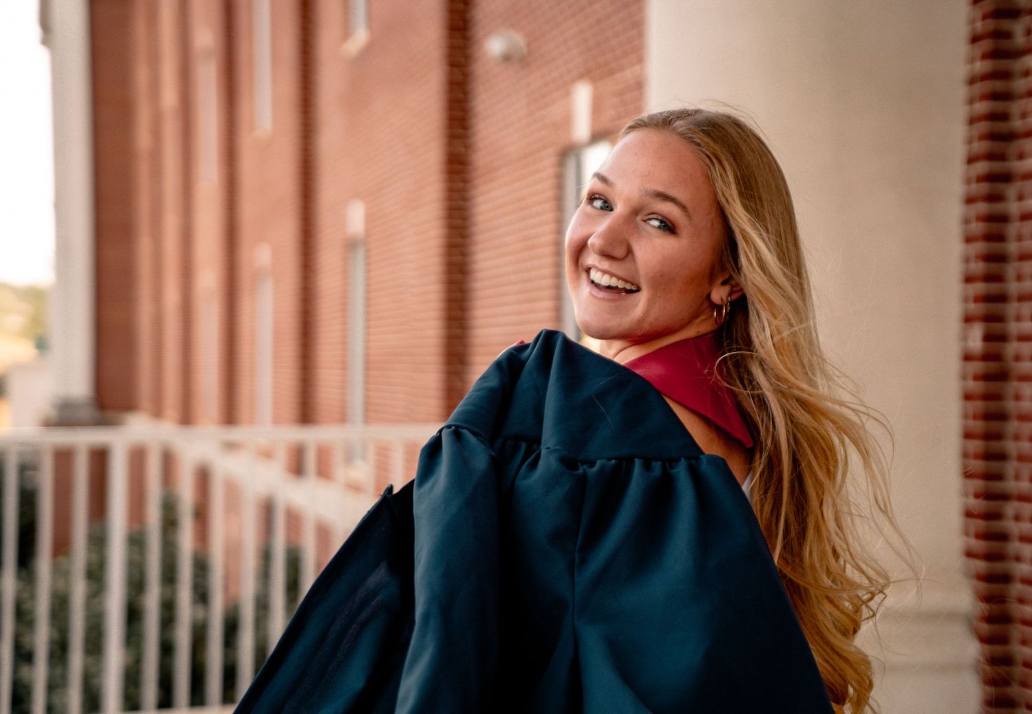 Smiling blonde woman in navy graduation gown with red hood poses outdoors near brick building.
