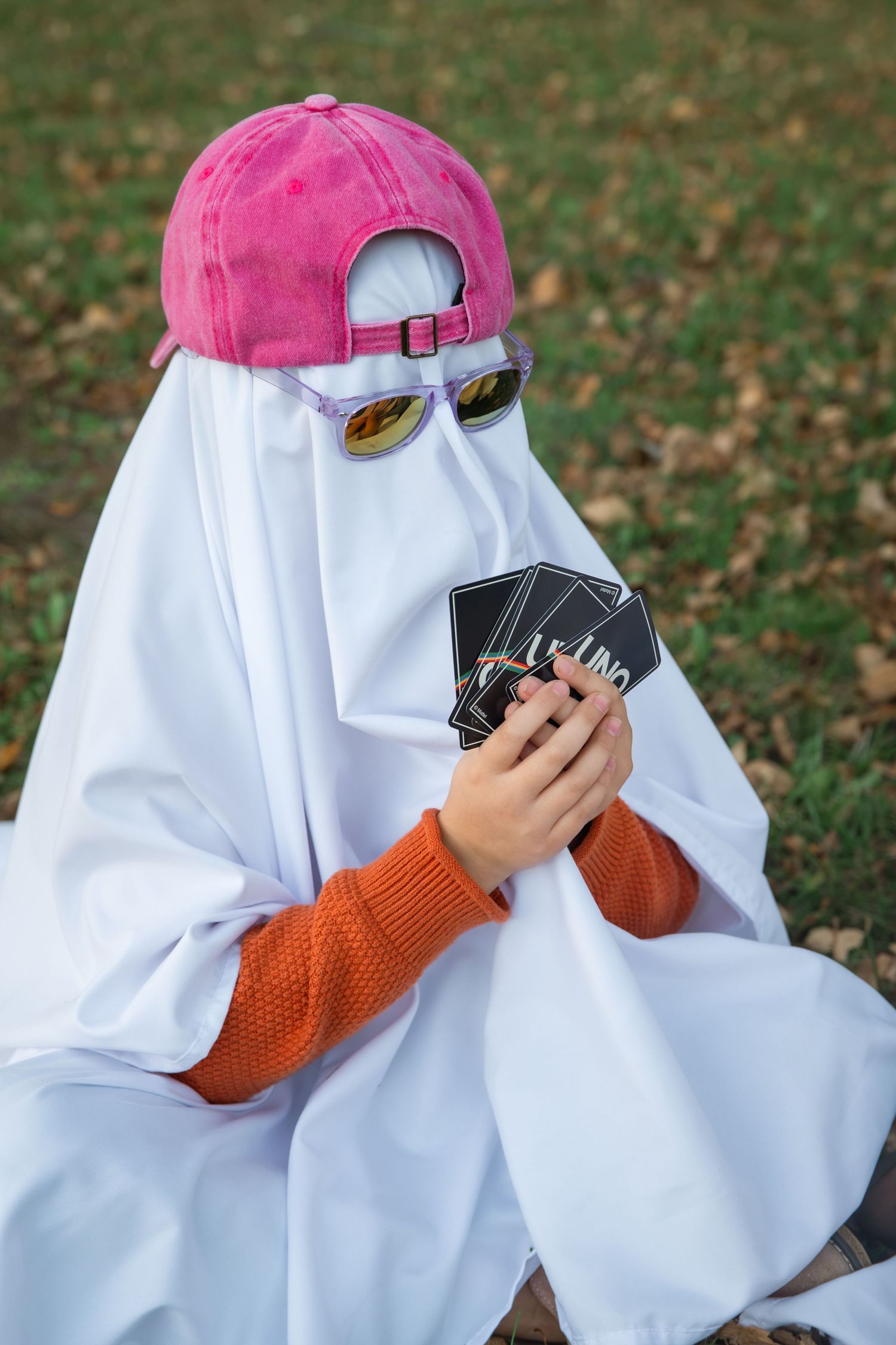Person in a white ghost sheet, pink cap and mirrored sunglasses sits on grass holding a hand of UNO cards.