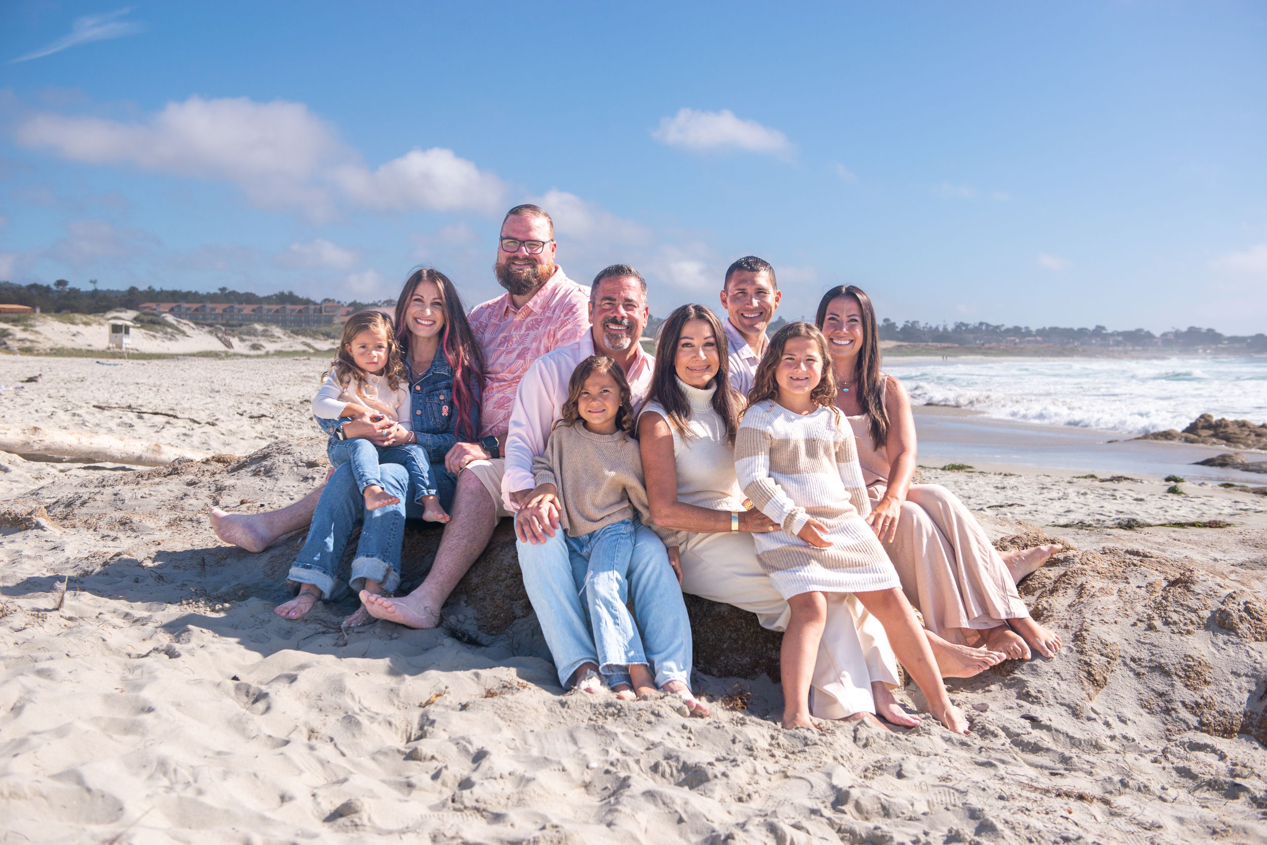 Smiling extended family of adults and children posing together on a sandy beach with ocean waves and blue sky in the background.