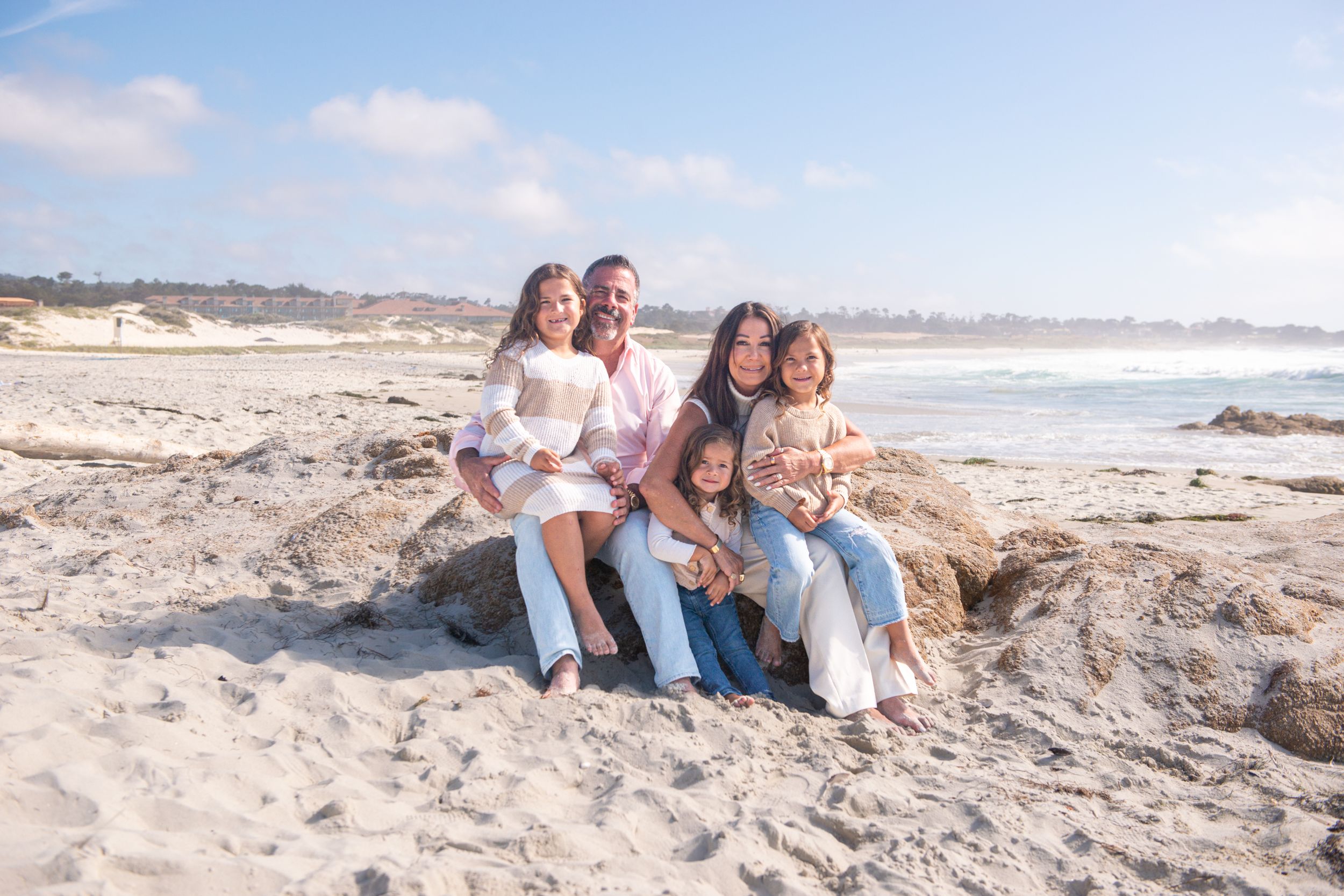 Smiling family of five sitting together on a rocky sandy beach near the ocean on a sunny day with blue sky.