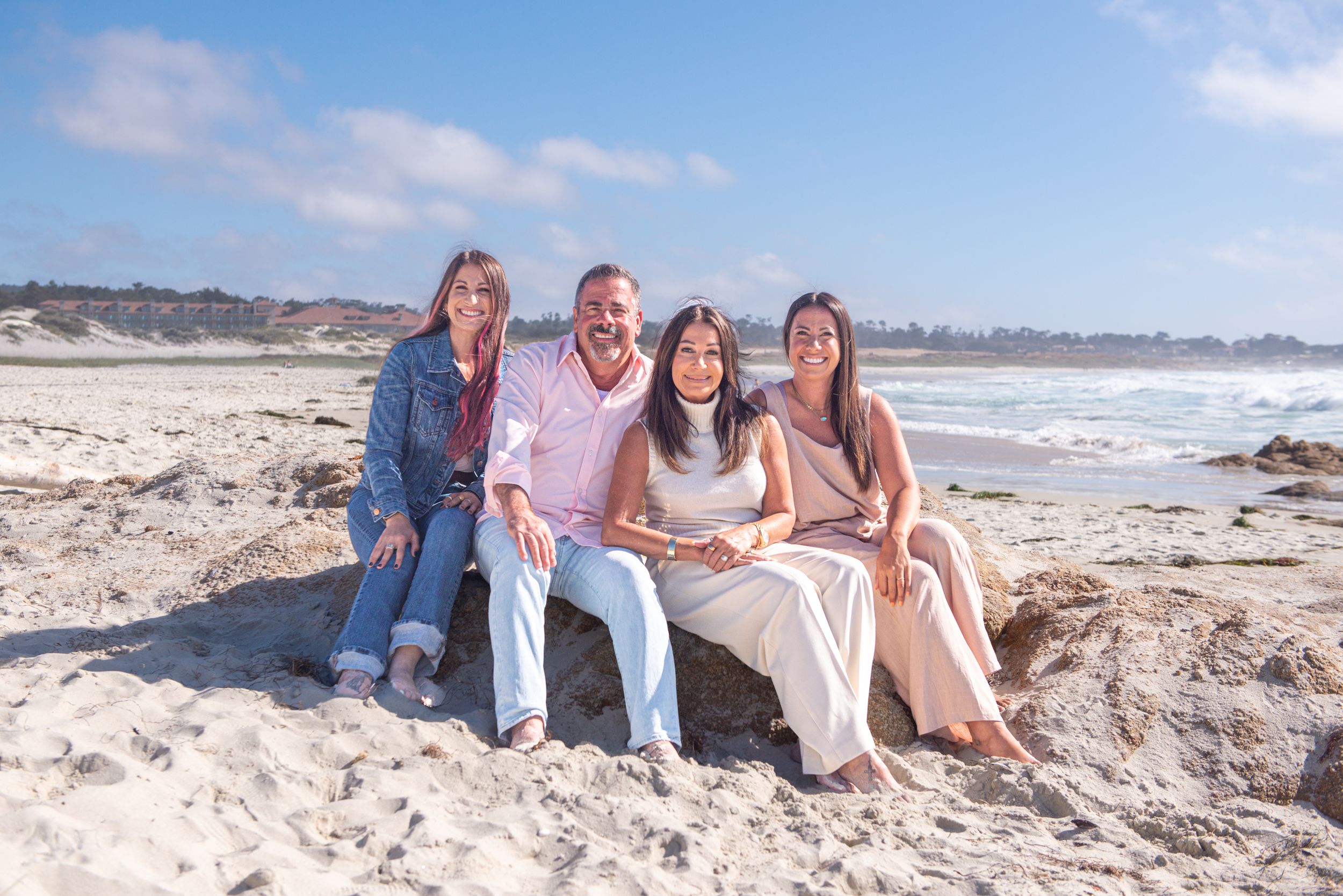 Smiling family of four sitting together on a sandy beach with rocks and ocean waves under a bright blue sky.