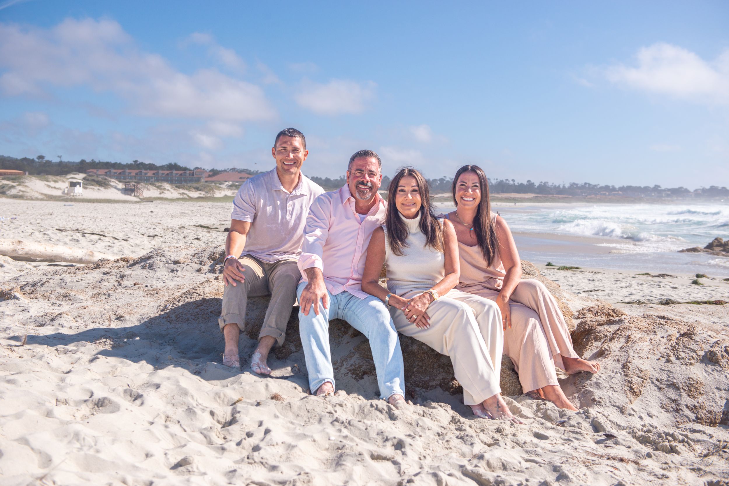Four family members in casual clothing sit close together on a sandy beach, smiling, with ocean waves and a blue sky behind them.
