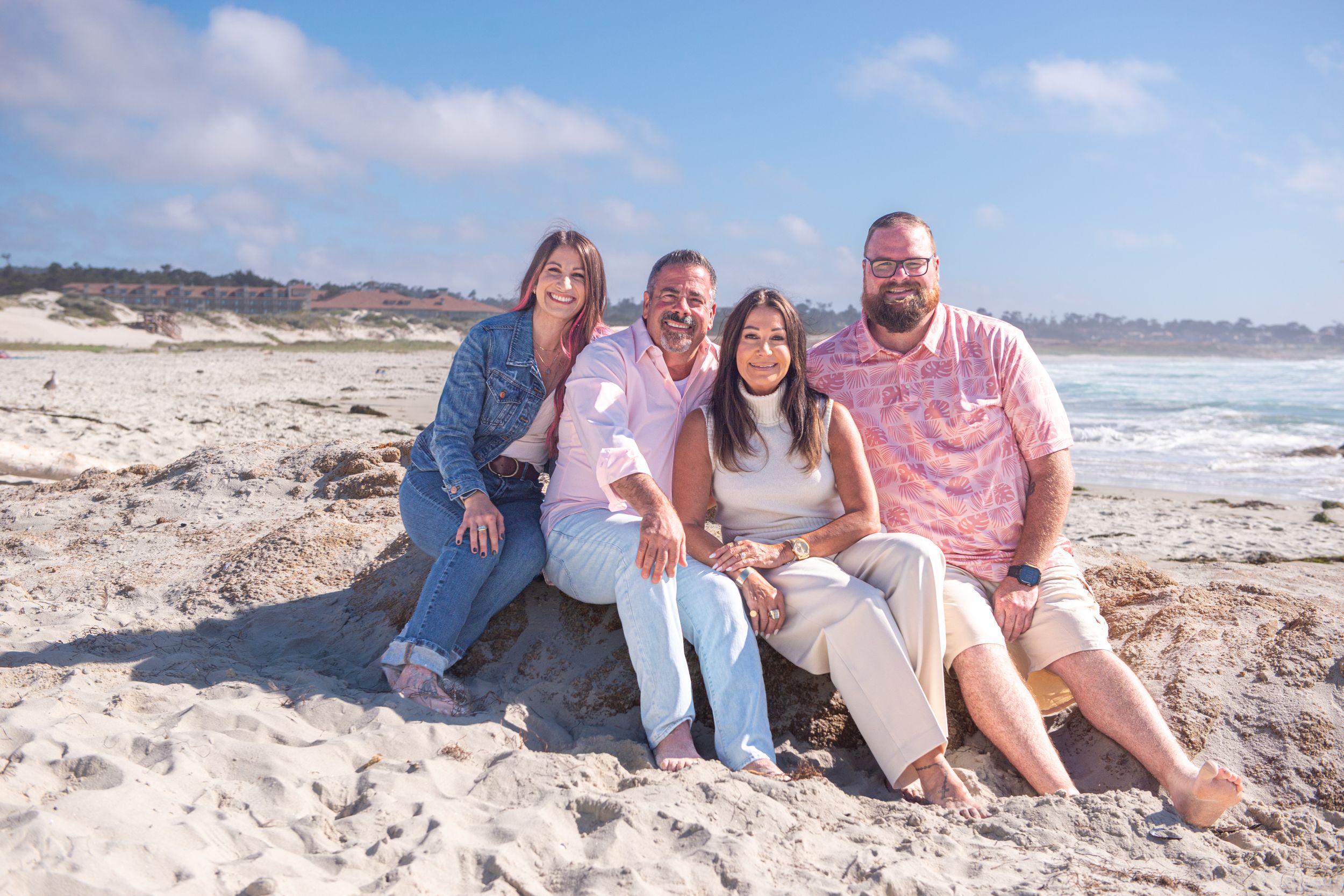 Four smiling adults in casual clothes sit together on a sandy rock at a beach with the ocean and blue sky behind them.