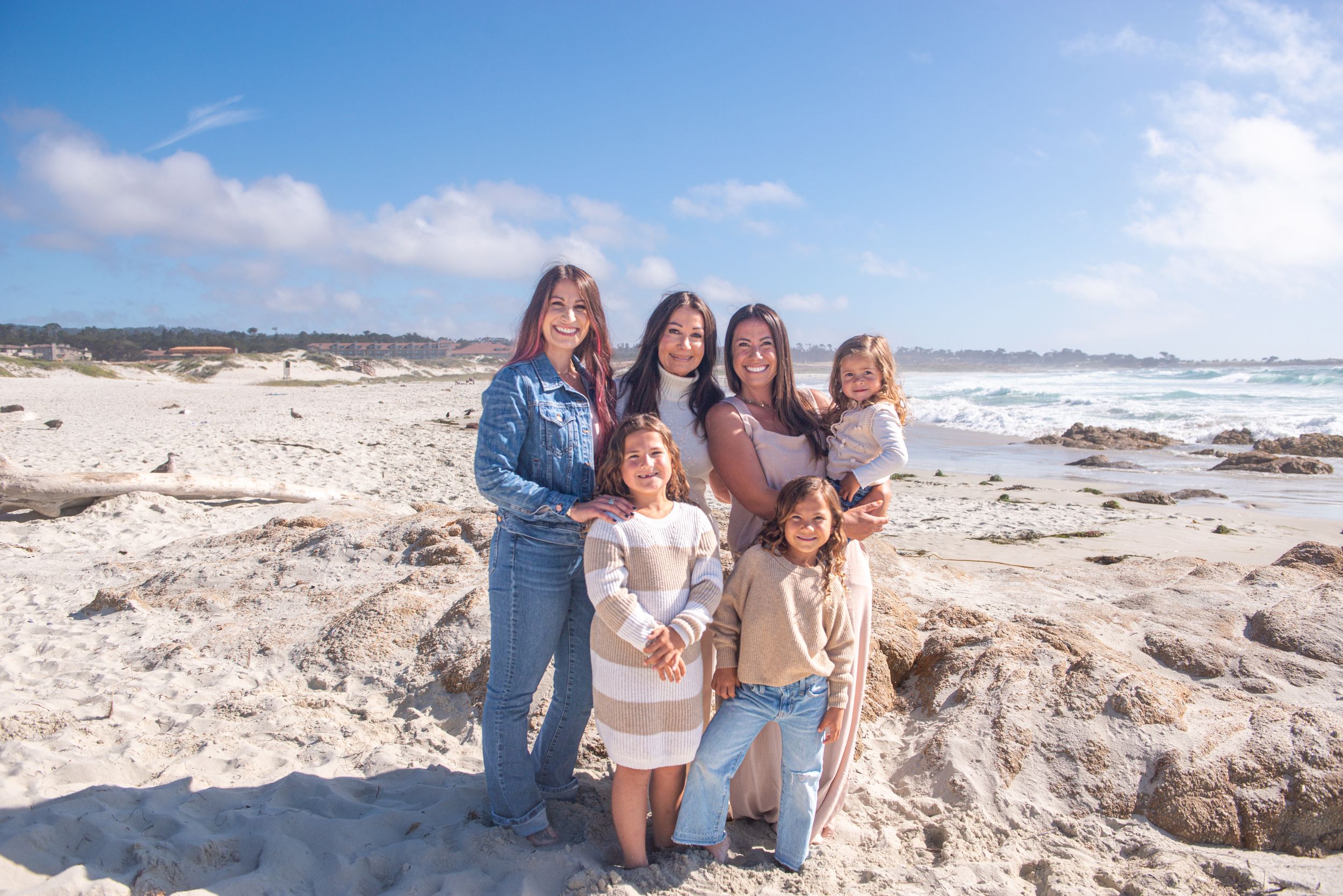Family of women and girls posing together on a sandy rocky beach with the ocean and blue sky behind them.
