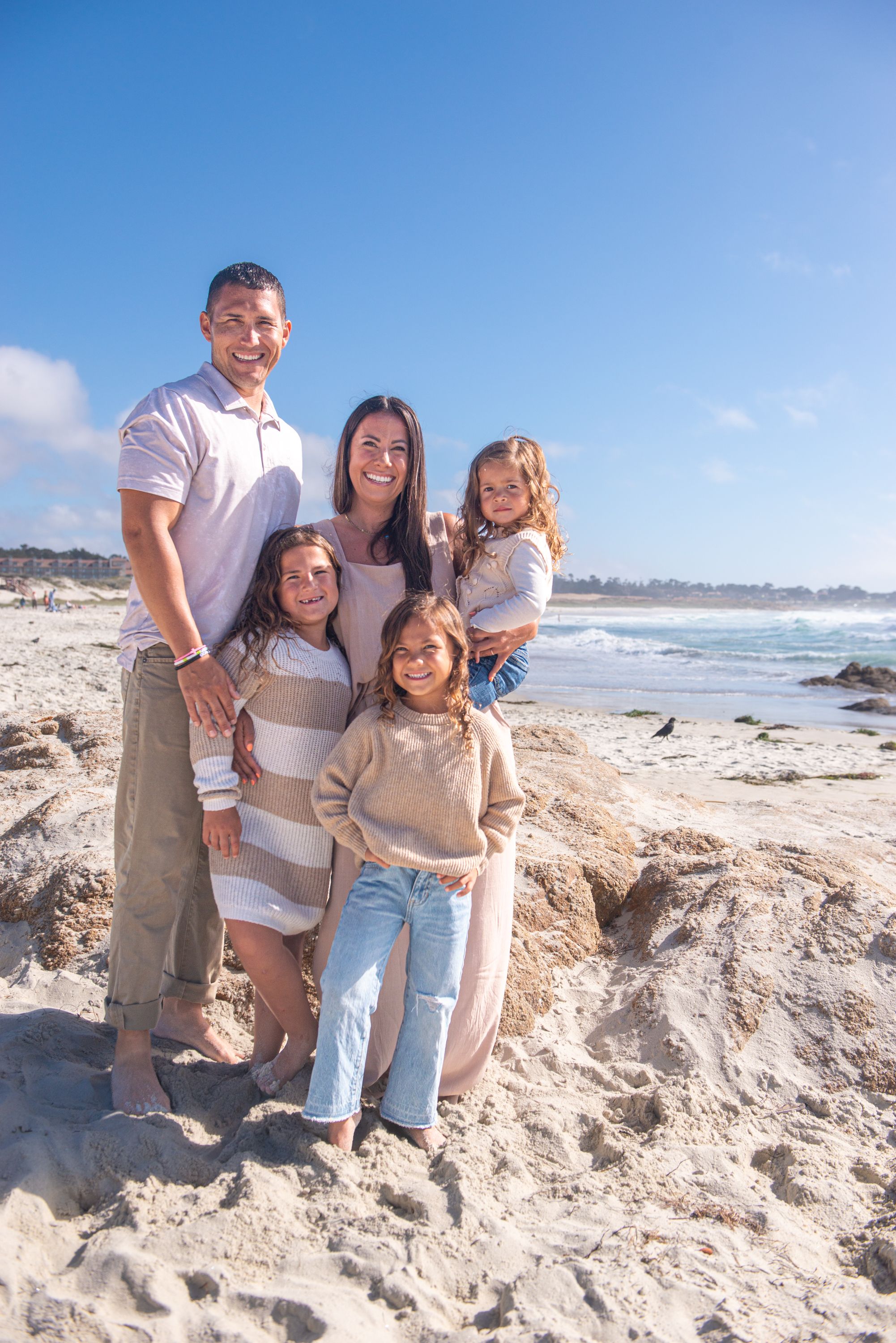 A smiling family of five poses together on a sandy rocky beach with ocean waves and a bright blue sky behind them.