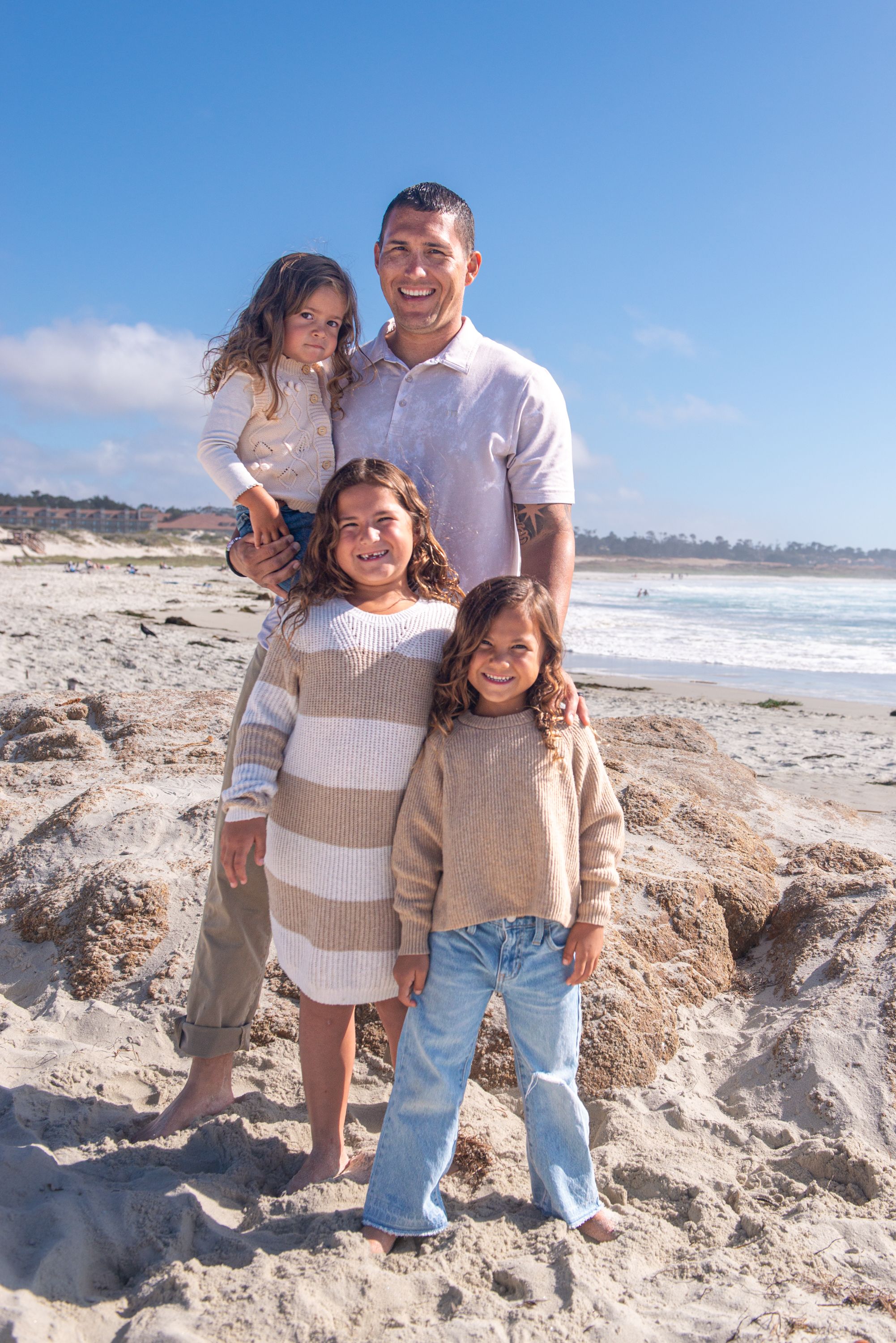 Smiling father and three young daughters in neutral outfits pose together on a sandy beach with ocean waves and blue sky behind them.
