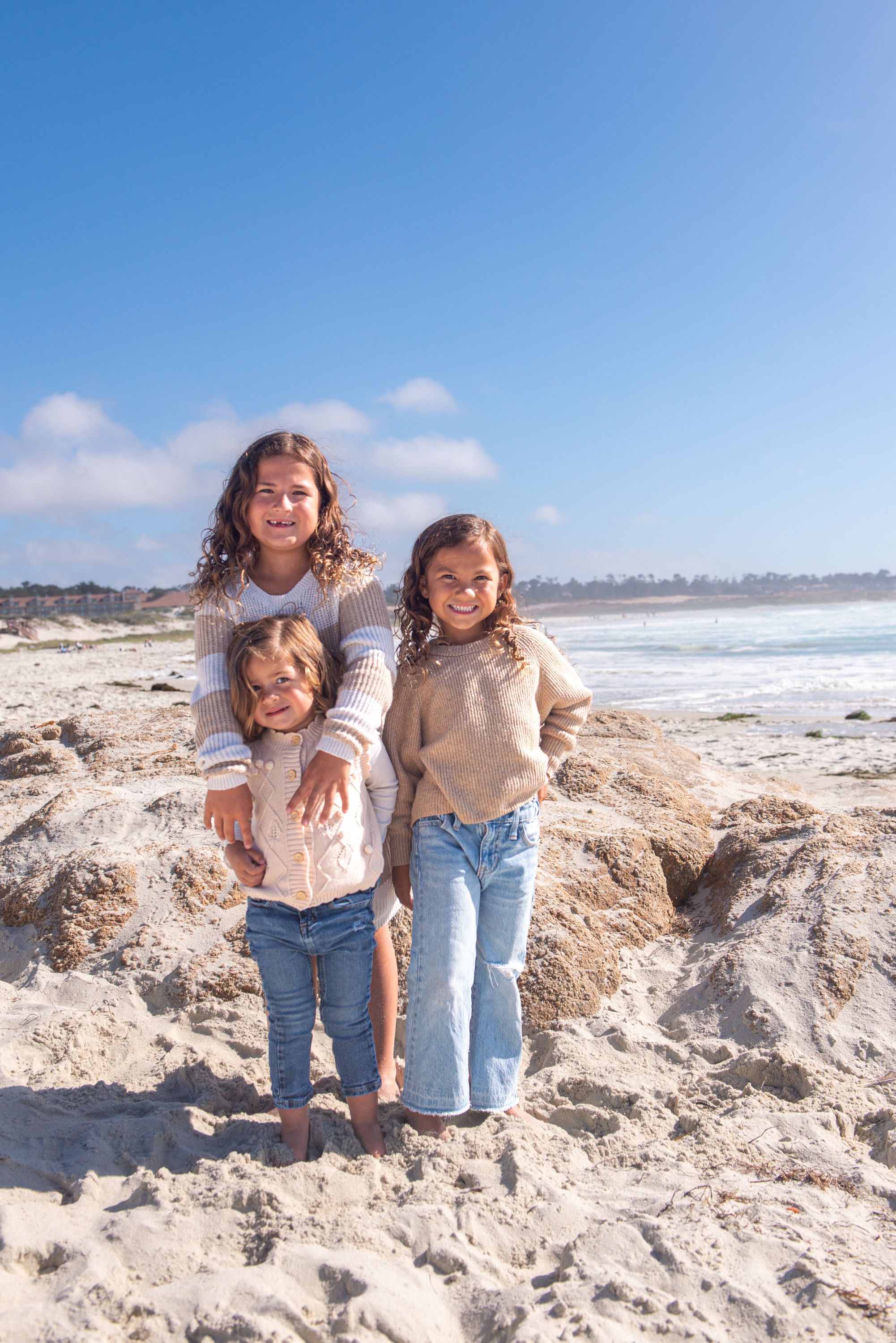 Three smiling young girls in sweaters and jeans pose together on a sandy beach with ocean waves and a clear blue sky behind them.