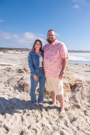 A smiling couple stands arm in arm on a sandy beach with ocean waves and a blue sky behind them.