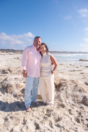 Smiling couple embracing on a sandy beach in light summer clothing with ocean waves and a blue sky in the background.