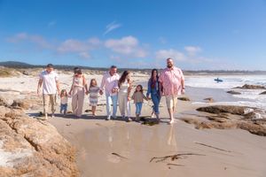 Large family of adults and children holding hands and walking together along a sandy beach with rocky shoreline under a blue sky.