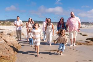 Multigenerational family walking and laughing together along a sandy beach with rocky shoreline under a bright blue sky.