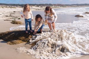 Three young girls in casual clothes explore a sandy beach, crouching and standing near rocks as foamy ocean waves wash ashore.