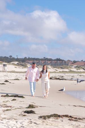 Smiling couple holding hands walking barefoot along a sandy beach with seagulls, seaweed, and coastal buildings under a blue sky.
