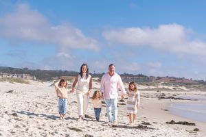 Family of five walking hand in hand along a sunny sandy beach with two adults and three young children near the shoreline.