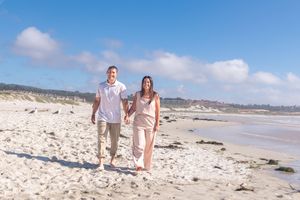 Smiling couple holding hands while walking barefoot along a sandy beach on a sunny day with blue sky and soft clouds.