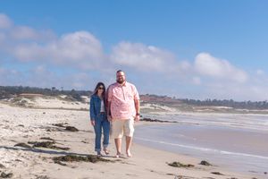 A couple walks hand in hand along a sandy shoreline on a sunny day with blue sky, soft clouds, and gentle ocean waves.
