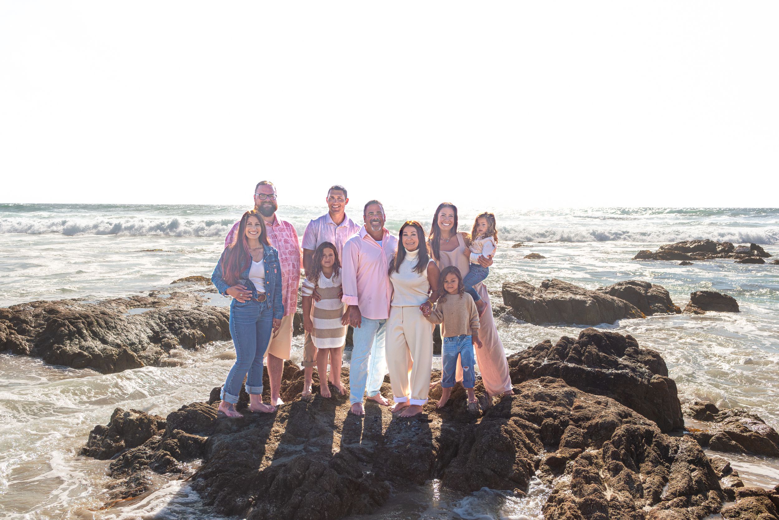 Large family in coordinated pink and neutral outfits poses together on dark rocks along the ocean shore at sunset.