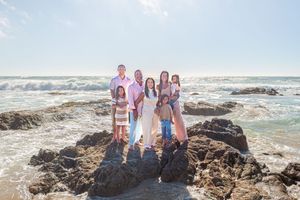 Family of six posing together on rocky coastal shoreline with ocean waves and bright blue sky in the background.
