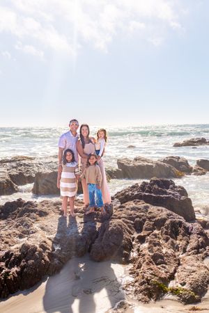 Family of five posing together on rugged coastal rocks with ocean waves and bright sunlit sky in the background.